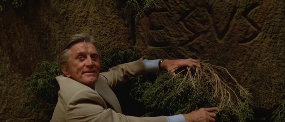 Kirk Douglas, açık renkli bir takım elbise içinde, yüzünde hafif bir şaşkınlık ifadesiyle görülüyor. Bir mağara veya antik kalıntının yosunlu taş duvarının önünde çömelmiş, sol eliyle bir çalılığı kenara çekerek, arkasındaki duvara kazınmış, kabaca oyulmuş ve sarmaşıklarla çevrili antik "IESUS" (muhtemelen "JESUS"un Latince yazımı) yazısını ortaya çıkarıyor. Işıklandırma, bu keşif anını ve Douglas'ın karakterinin bu eski sembolle yüzleşmesini dramatik bir şekilde aydınlatıyor.